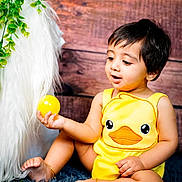 Aithen is registered to the contest to win money with this photo: baby, child, yellow_outfit, duck_design, ball, curious, sitting, blue_rug, furry_texture, wooden_background, green_leaves, indoors, cute, happy, playful, barefoot, anklet, portrait, infant, toy