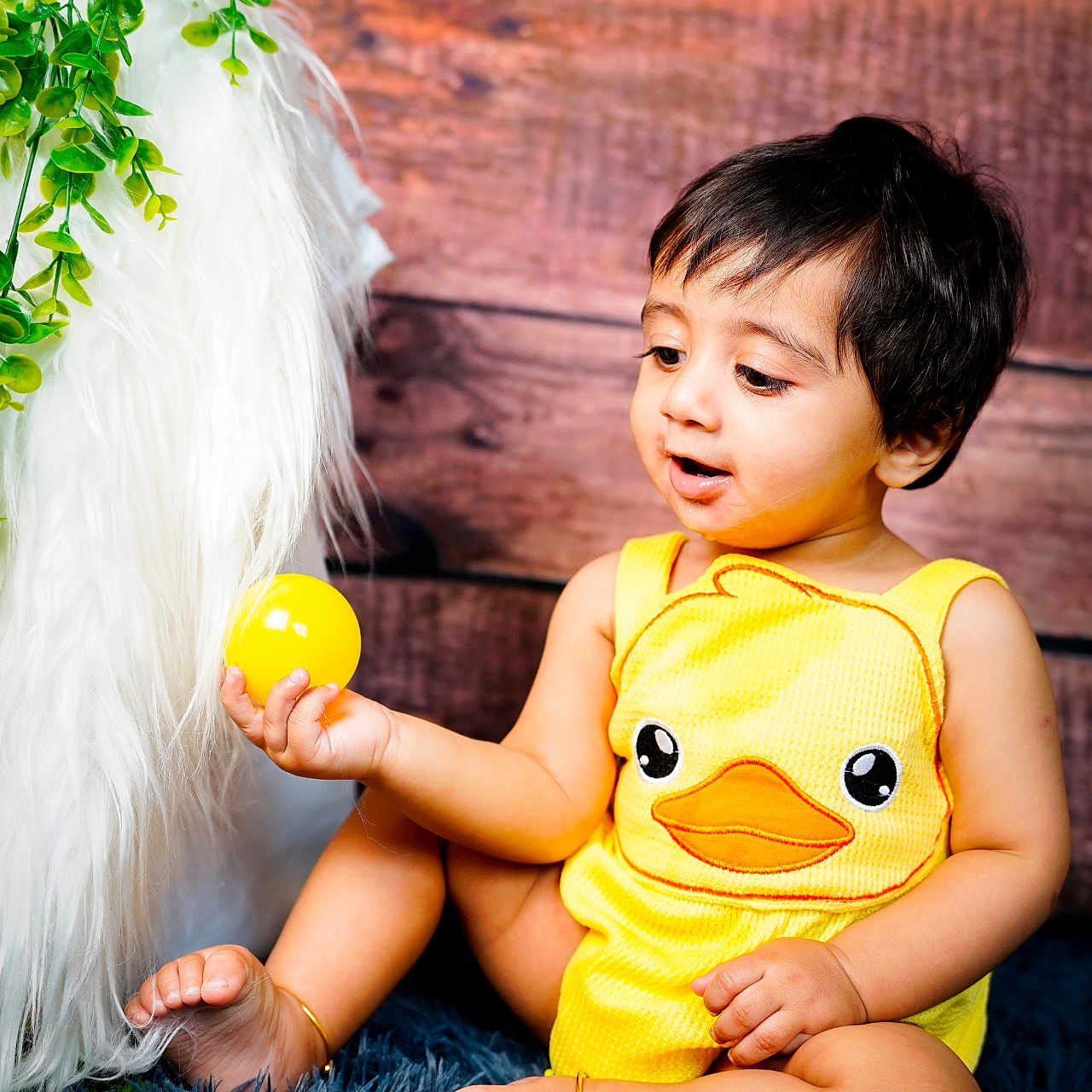 Aithen is registered to the contest to win money with this photo: anklet, baby, ball, barefoot, blue_rug, child, curious, cute, duck_design, furry_texture, green_leaves, happy, indoors, infant, playful, portrait, sitting, toy, wooden_background, yellow_outfit