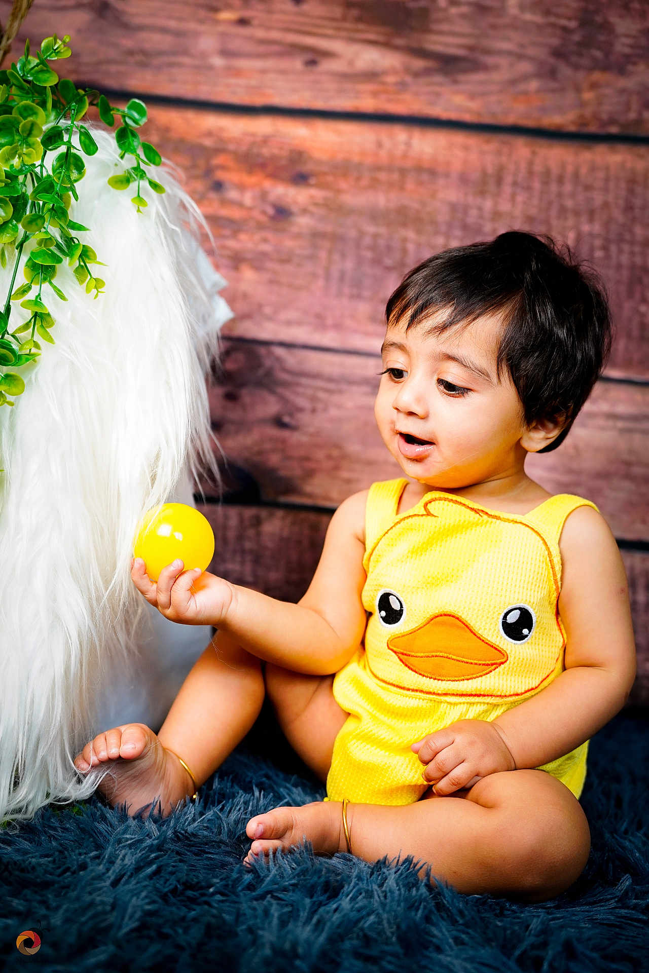 Aithen is registered to the contest to win money with this photo: baby, child, yellow_outfit, duck_design, ball, curious, sitting, blue_rug, furry_texture, wooden_background, green_leaves, indoors, cute, happy, playful, barefoot, anklet, portrait, infant, toy