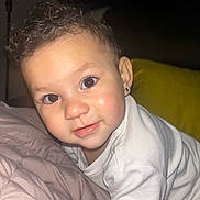 Skye is registered to the contest to win money with this photo: baby, child, curly_hair, face, eyes, white_outfit, earring, indoor, pillow, soft_surface, close_up, portrait, cute, smiling, infant, person, skin, head, young, cozy