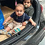 toddler, baby, child, car_trunk, pacifier, striped_clothing, shorts, sneakers, sandals, blue_bag, pavement, curious, sitting, holding, black_car, outdoor, expression, candid, cute, two_children