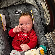 Malachi is registered to the contest to win money with this photo: baby, child, stroller, shopping_cart, red_shirt, stuffed_toy, blanket, indoor, cute, smiling, infant, clothing, face, hand, toy, seat, metal, comfort, person, casual