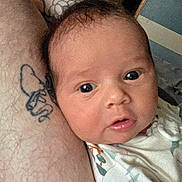 Cooper is registered to the contest to win money with this photo: baby, infant, close_up, face, eyes, skin, tattoo, arm, person, indoor, soft_lighting, curious, newborn, clothing, portrait, human, head, expression, cuddle, parent