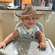 Reshard Mcelroy Jr is registered to the contest to win money with this photo: baby, child, cowboy_hat, high_chair, smile, happy, indoor, kitchen, wooden_cabinets, feeding_chair, infant, person, cute, adorable, straps, table, face, expression, home, decor