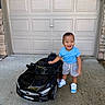 toddler, child, smiling, toy_car, black_car, outdoor, garage_door, concrete, sneakers, shorts, blue_shirt, happy, standing, play, vehicle, daylight, person, cute, small_car, front_view