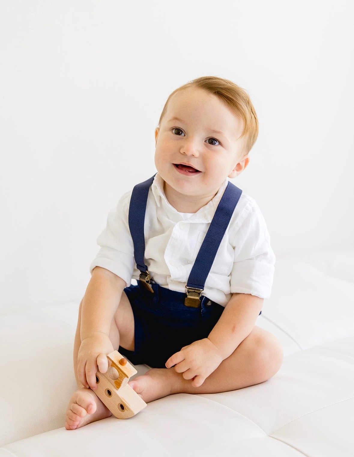 Rocky joined the competition — help win amazing prizes! toddler, child, smiling, white_shirt, navy_suspender, wooden_toy, toy_ship, barefoot, sitting, cross_legged, light_background, indoor, cute, happy, young_child, playful, portrait, innocence, minimalist, baby