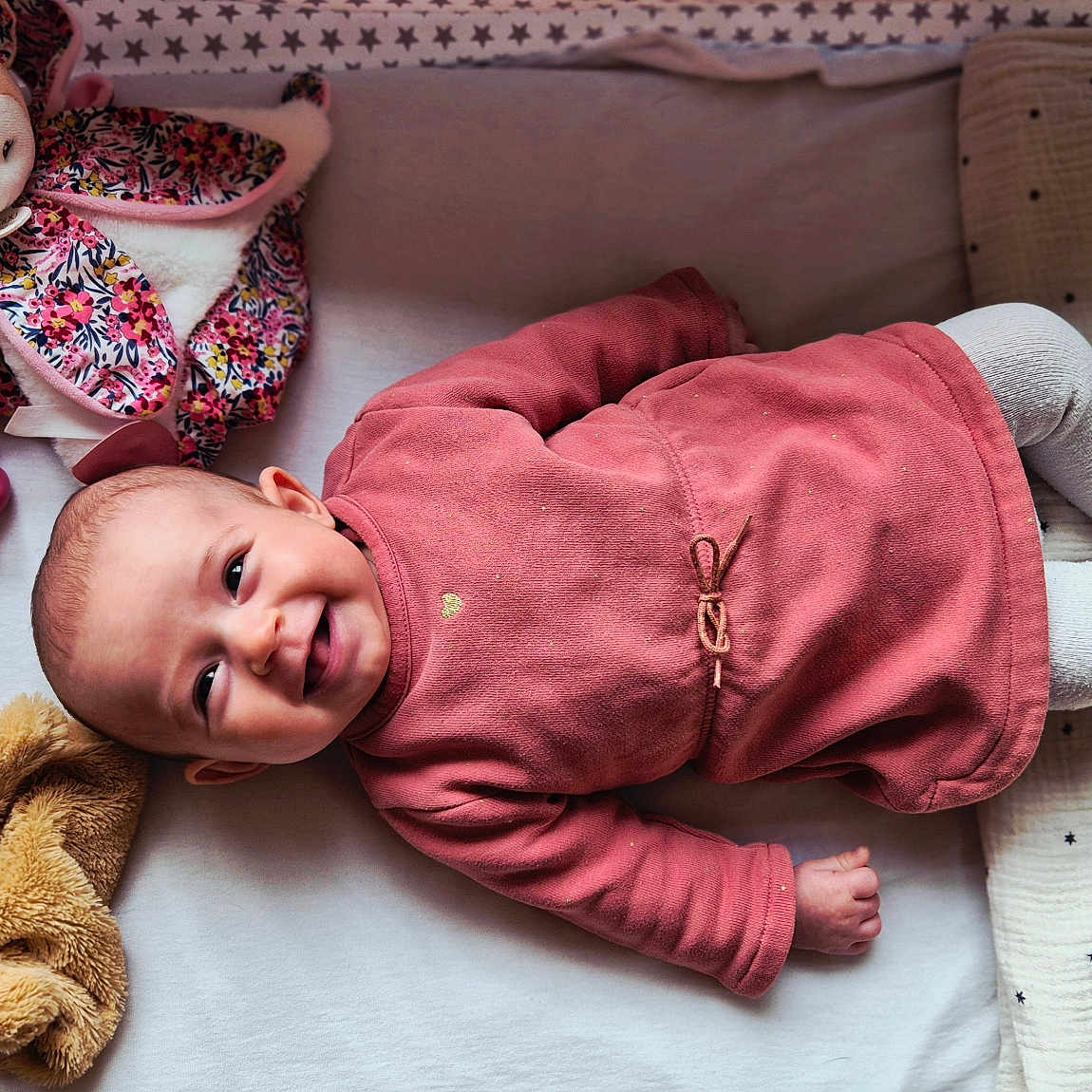 Louisa a rejoint le concours — aidez-le/la à gagner de superbes lots ! baby, bedding, blanket, child, cozy, cute, face, happy, head, indoors, infant, lying_down, pacifier, pink_clothing, sleep_area, smiling, soft_toys, stars_pattern, stuffed_animal, toddler