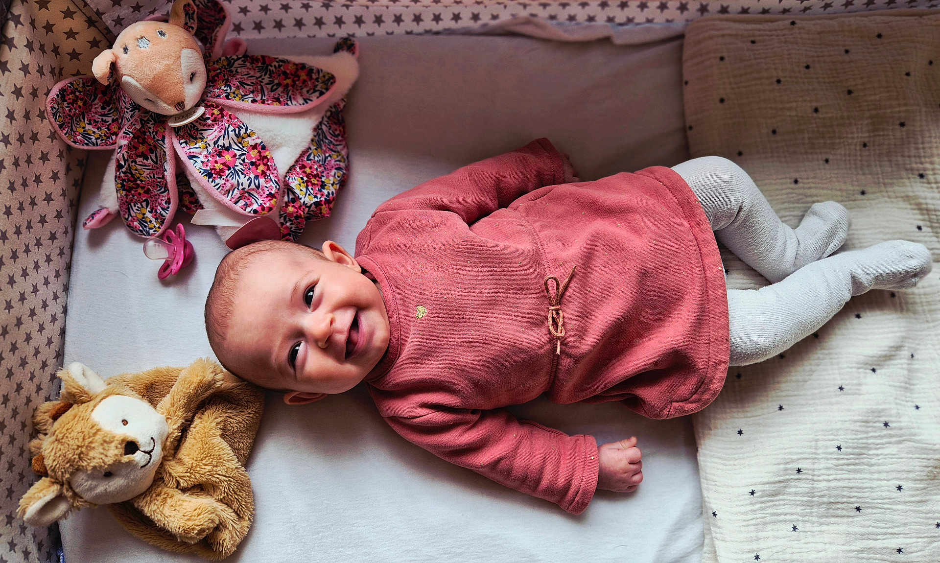 Louisa a rejoint le concours — aidez-le/la à gagner de superbes lots ! baby, infant, smiling, lying_down, pink_clothing, stuffed_animal, pacifier, bedding, stars_pattern, toddler, cute, child, soft_toys, blanket, indoors, happy, face, head, sleep_area, cozy