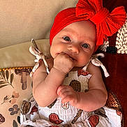 Poppy is registered to the contest to win money with this photo: baby, infant, red_headband, bow, dress, patterned_clothing, hand_in_mouth, cute, child, cushion, floral_pattern, seated, indoor, portrait, closeup, smiling, happy, skin, face, person