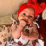 baby, infant, red_headband, bow, dress, patterned_clothing, hand_in_mouth, cute, child, cushion, floral_pattern, seated, indoor, portrait, closeup, smiling, happy, skin, face, person
