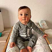 Tylio participe au concours pour gagner de l'argent avec cette photo : baby, child, infant, cute, rosy_cheeks, grey_outfit, sitting, changing_pad, floral_pattern, wooden_surface, indoor, curious, calm, small_feet, hands, face, eyes, hair, wall, home