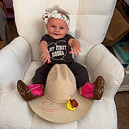 Arabella is registered to the contest to win money with this photo: baby, headband, cowboy_boots, cowboy_hat, rubber_duck, white_chair, smiling, shirt, infant, cute, indoors, child, sitting, plush_chair, clothing, footwear, toy, happy, portrait, accessory