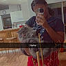 person, cat, mirror_selfie, phone, leopard_print, red_pants, home_interior, kitchen, wood_floor, decorative_sticks, berries, long_hair, braids, gray_cat, tongue_out, furniture, chair, table, casual_clothing, reflection