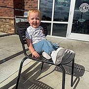 Asher is registered to the contest to win money with this photo: blonde_hair, brick_wall, building, casual_clothing, chair, child, daylight, denim_pants, glass_door, happy, metal_chair, outdoor, reflection, shadow, short_sleeve_shirt, sidewalk, smiling, sunlight, toddler, white_shoes