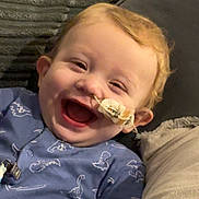 Arlo-Carl joined the competition — help win amazing prizes! baby, child, smiling, laughing, indoors, onesie, dinosaur_pattern, couch, pillow, medical_tube, happy, infant, face, head, person, clothing, cozy, young_child, cute, relaxed