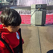 Mateo is registered to the contest to win money with this photo: child, toddler, fountain, water, pink_water, outdoor, city, architecture, building, stairs, balustrade, sign, chain, pavement, red_clothing, jacket, dark_hair, portrait, tourist_attraction, daylight
