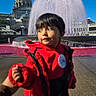 child, toddler, red_coat, outdoor, fountain, water, domed_building, architecture, city, landmark, travel, tourism, portrait, face, dark_hair, looking_away, winter_clothing, scarf, daylight, blue_sky