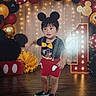 balloons, birthday, black_shoes, bow_tie, celebration, child, colorful_background, costume, decorations, indoors, marquee_lights, mickey_mouse, number_one, party, portrait, red_shorts, smiling, standing, toddler, wooden_floor