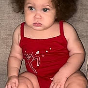 Jalaya joined the competition — help win amazing prizes! baby, child, curly_hair, red_bow, red_outfit, socks, couch, sitting, indoor, cute, serious_expression, chubby_arms, chubby_legs, infant, portrait, face, person, skin, furniture, home