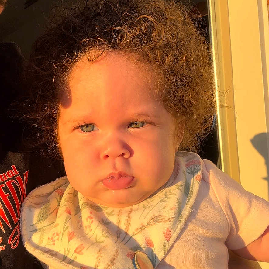 Jalaya is registered to the contest to win money with this photo: baby, bib, cheeks, child, clothing, curly_hair, cute, doorway, expression, face, holding, indoors, infant, light, person, portrait, serious, skin, sunlight, young