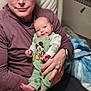 baby, man, glasses, chair, clothing, baby_clothes, mickey_mouse, smile, person, indoor, cushion, blanket, hand, face, seated, watch, ring, hoodie, relaxation, parenting