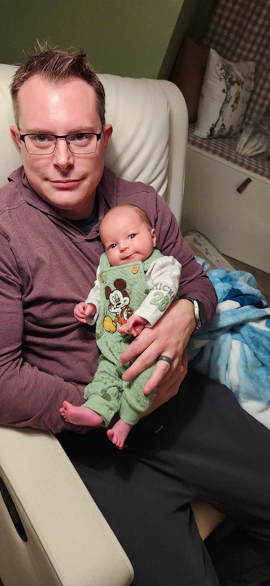 Ryan is registered to the contest to win money with this photo: baby, man, glasses, chair, clothing, baby_clothes, mickey_mouse, smile, person, indoor, cushion, blanket, hand, face, seated, watch, ring, hoodie, relaxation, parenting