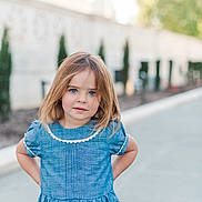 Camille joined the competition — help win amazing prizes! child, girl, blue_dress, blue_eyes, outdoor, portrait, standing, curious, pathway, greenery, wall, daylight, young, casual_clothing, person, cute, innocent, natural_light, blonde_hair, short_sleeves