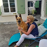 Loona participe au concours pour gagner de l'argent avec cette photo : woman, dog, german_shepherd, chair, outdoor, smile, pet, relaxing, courtyard, concrete, window, green_shutters, footwear, barefoot, casual_clothing, glasses, sunlight, fence, door, plants