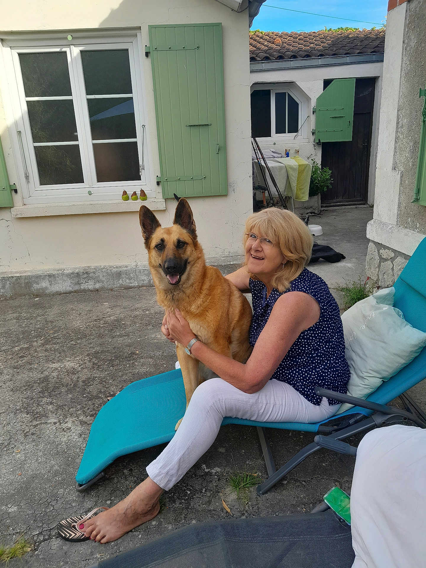 Loona participe au concours pour gagner de l'argent avec cette photo : woman, dog, german_shepherd, chair, outdoor, smile, pet, relaxing, courtyard, concrete, window, green_shutters, footwear, barefoot, casual_clothing, glasses, sunlight, fence, door, plants