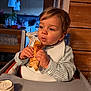 toddler, child, high_chair, food, wrap, eating, indoor, kitchen, bib, clothing, table, face, hands, curly_hair, young_child, focused, meal_time, wood_paneling, soft_lighting, casual