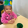 child, smiling, crown, handmade, colorful, pink_jacket, table, green_cup, indoor, classroom, young_child, seated, face, happy, playful, kids, craft, portrait, person, casual