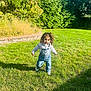 child, toddler, grass, outdoor, sunlight, greenery, overalls, curly_hair, walking, lawn, brick_wall, hedge, casual_clothing, daylight, nature, smiling, playful, person, young_child, park