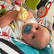 Gabriel participe au concours pour gagner de l'argent avec cette photo : baby, pacifier, toy, play_mat, infant, colorful, soft_toy, child, cute, indoor, lying_down, curious, blue, fabric, pattern, hand, face, head, eyes, smile