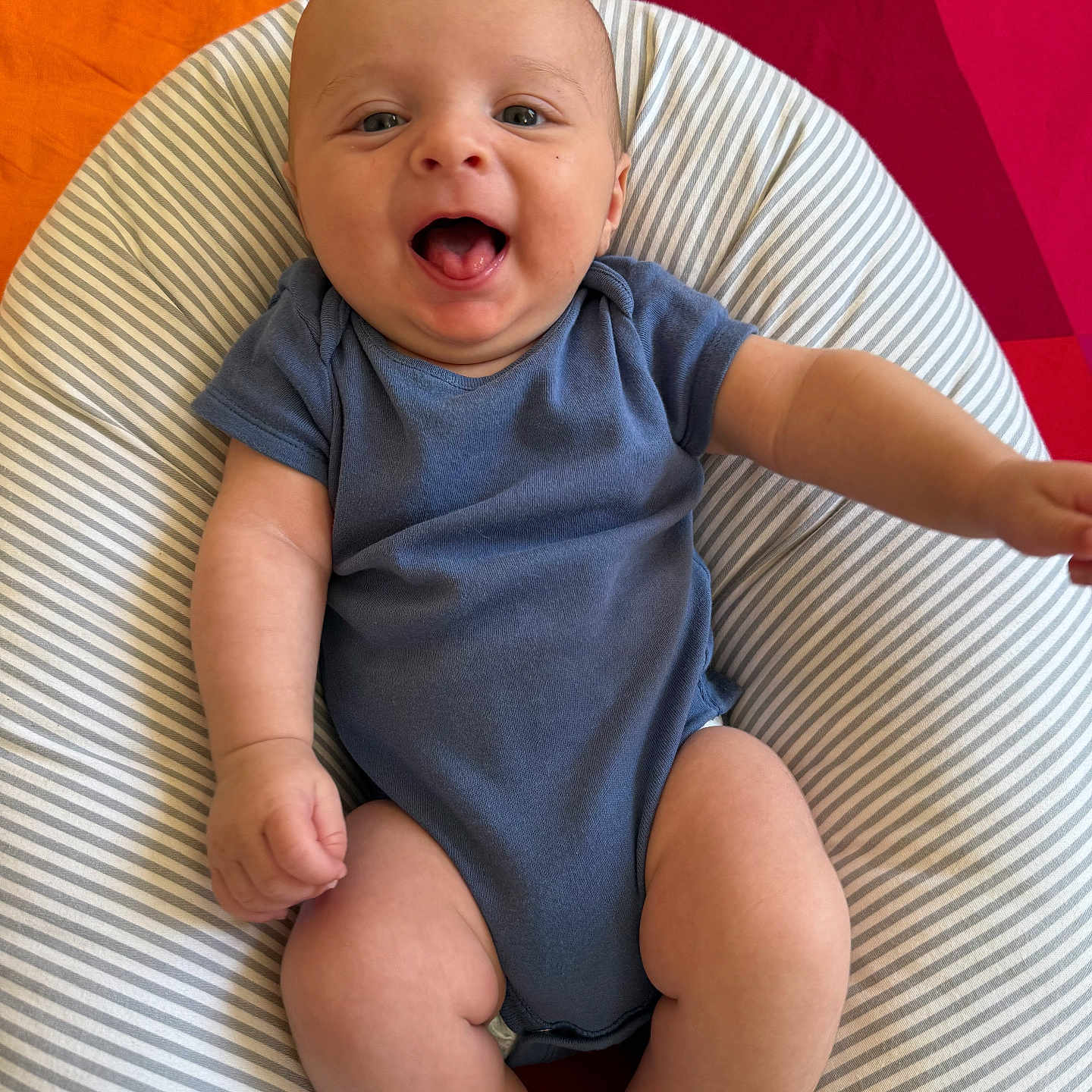Gabriel participe au concours pour gagner de l'argent avec cette photo : arms, baby, blue_onesie, child, chubby_legs, cute, face, happy, indoor, infant, mouth_open, orange_background, person, portrait, red_background, sitting, skin, smiling, striped_cushion, young_child