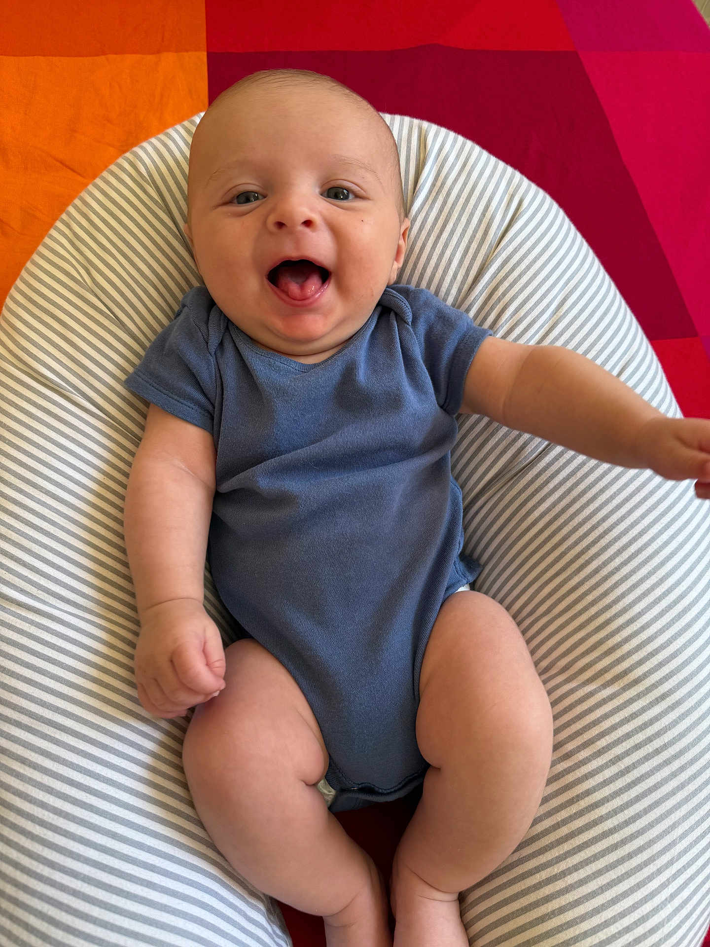 Gabriel participe au concours pour gagner de l'argent avec cette photo : baby, infant, smiling, blue_onesie, striped_cushion, red_background, orange_background, happy, cute, child, portrait, skin, chubby_legs, arms, face, mouth_open, indoor, person, sitting, young_child