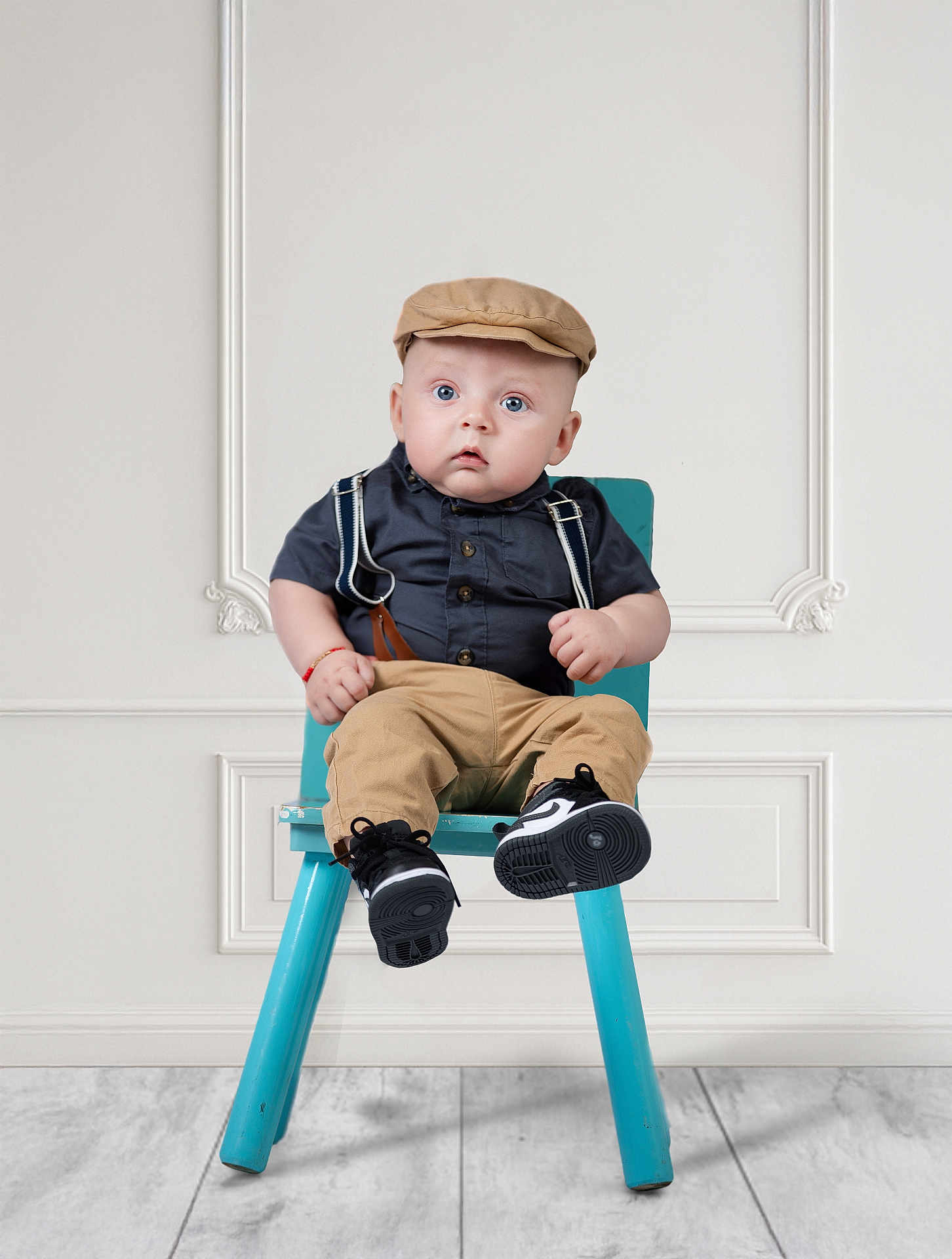 Victor is registered to the contest to win money with this photo: baby, child, blue_eyes, suspenders, khaki_pants, cap, chair, footwear, black_shoes, indoor, wood_floor, white_wall, expression, portrait, sitting, fashion, cute, young_child, person, studio
