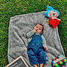 baby, child, infant, outdoor, grass, blanket, denim_overalls, plaid_shirt, plush_toy, clown_doll, colorful_toy, letter_board, portrait, lying_down, looking_at_camera, hand_to_mouth, monthly_milestone, natural_light, greenery, family_photo