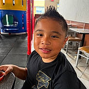 George joined the competition — help win amazing prizes! child, boy, smiling, portrait, restaurant, indoors, play_area, playground, table, chair, food, dipping_sauce, black_shirt, spiky_hair, seated, happy, face, person, colorful_background, casual