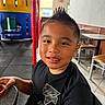 child, boy, smiling, portrait, restaurant, indoors, play_area, playground, table, chair, food, dipping_sauce, black_shirt, spiky_hair, seated, happy, face, person, colorful_background, casual