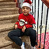 George is registered to the contest to win money with this photo: child, boy, christmas, santa_hat, festive_sweater, red_clothing, staircase, indoor, portrait, sitting, smiling, face, railing, carpet, wood_floor, holiday, decorations, socks, home, seasonal