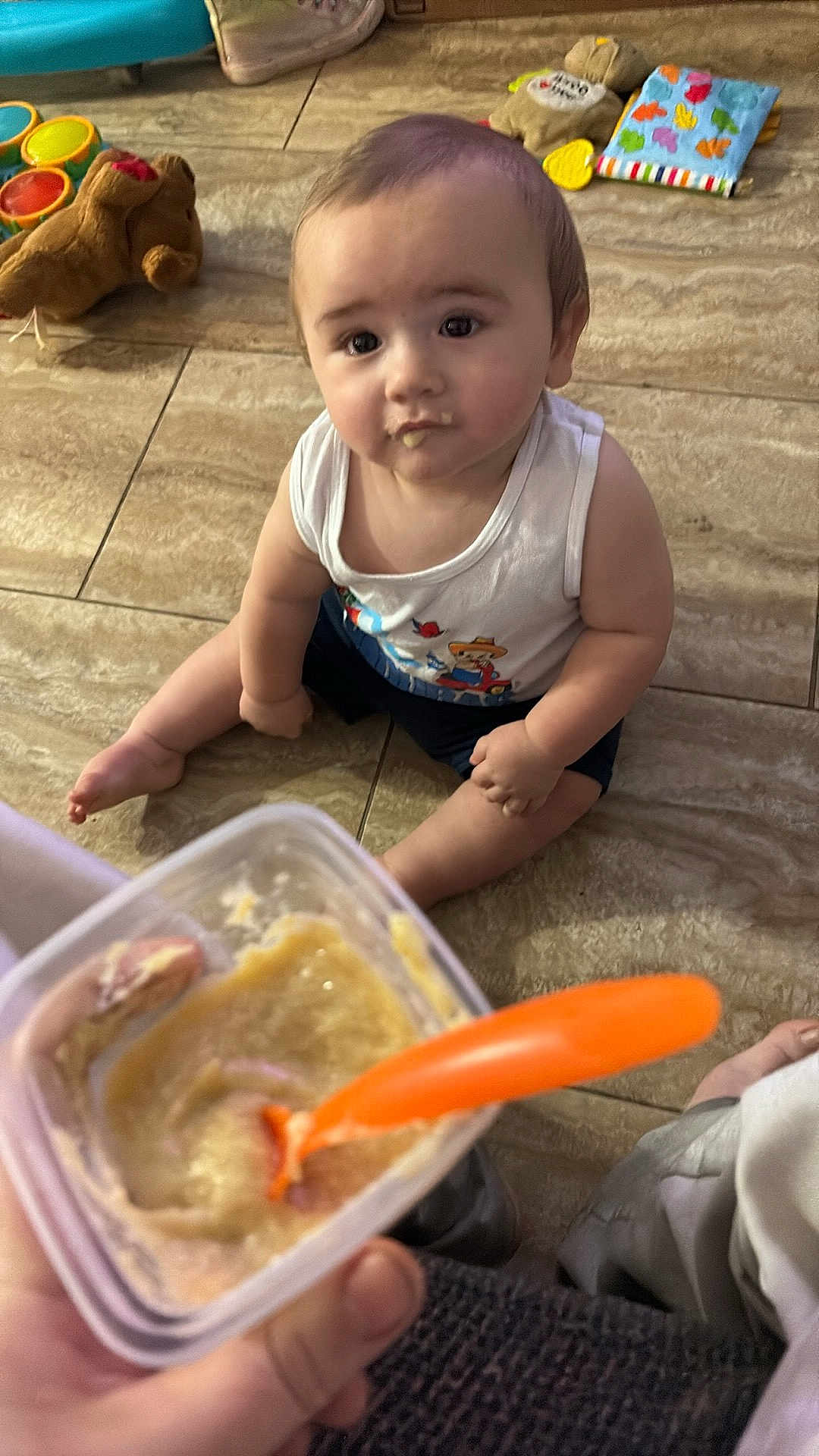 Zaidyn joined the competition — help win amazing prizes! baby, child, food, baby_food, spoon, floor, tile, toy, cushion, hand, container, clothing, sitting, indoor, feeding, curious, face, mouth, eyes, person