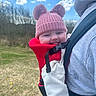 Olive is registered to the contest to win money with this photo: baby, child, smiling, hat, pink_hat, pom_poms, baby_carrier, red_carrier, hoodie, grey_hoodie, outdoor, sky, clouds, grass, field, person, happy, cute, portrait, daytime