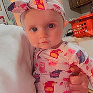 Charlotte is registered to the contest to win money with this photo: baby, child, blue_eyes, headband, onesie, owl_pattern, hand, person, indoor, kitchen, laundry_basket, orange_basket, cabinet, doorway, curtain, floor_mat, human, portrait, closeup, cute