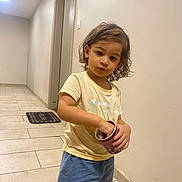Soane participe au concours pour gagner de l'argent avec cette photo : child, kid, curly_hair, yellow_tshirt, blue_shorts, snack, container, hallway, tile_floor, door, mat, indoor, person, young_child, casual_clothing, standing, looking_at_camera, hand_in_container, light, wall