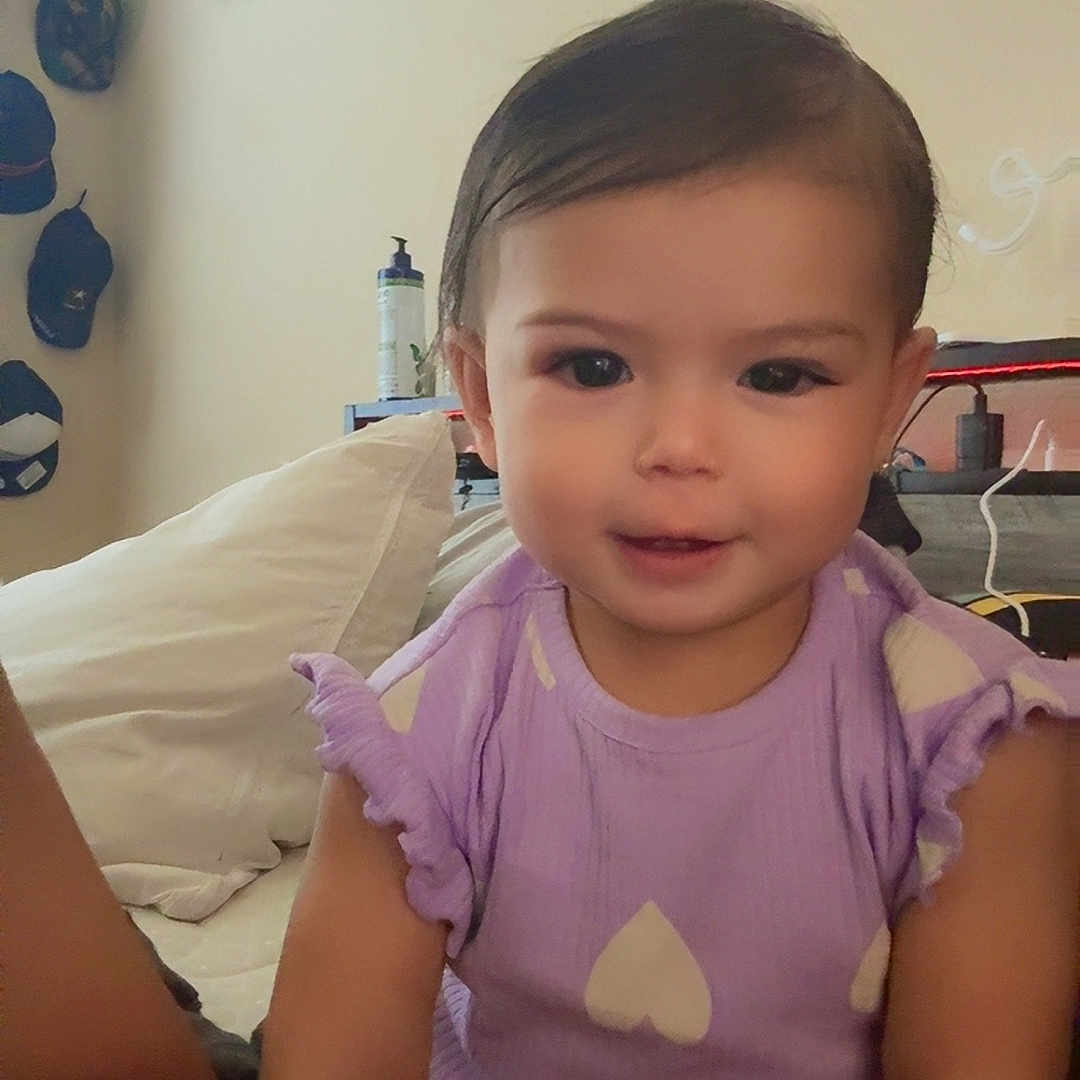 Skylar is registered to the contest to win money with this photo: toddler, child, purple_onesie, heart_pattern, smiling, indoor, bed, pillow, caps_on_wall, framed_certificate, person, face, hair, cute, comfortable, room, casual, young_child, seated, soft_lighting