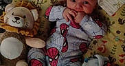 Mateo is registered to the contest to win money with this photo: baby, infant, spiderman_onesie, plush_toy, lion_toy, blanket, pillow, patterned_fabric, child, cute, indoor, portrait, baby_clothing, soft_toy, lying_down, hand_to_mouth, curious, face, baby_bedding, cozy