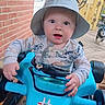 baby, child, hat, toy_car, dinosaur_print, blue, steering_wheel, outdoor, wooden_deck, brick_wall, dog, pet, fence, decorative_star, clothing, person, portrait, smiling, face, hands