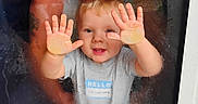 Mattie is registered to the contest to win money with this photo: toddler, child, baby, face, smiling, hands, palms, window, glass, raindrops, reflection, indoor, portrait, sitting, gray_shirt, blond_hair, blue_text, home, candid, playful
