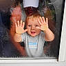 baby, blond_hair, blue_text, candid, child, face, glass, gray_shirt, hands, home, indoor, palms, playful, portrait, raindrops, reflection, sitting, smiling, toddler, window