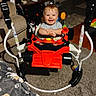 Mattie is registered to the contest to win money with this photo: baby, child, smiling, happy, indoor, living_room, activity_walker, toy, red, portrait, sitting, standing, face, eyes, hair, carpet, couch, blanket, furniture, home